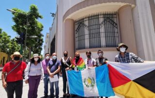 A group of individuals stand in the sun in front of a building in Guatemala holding the Guatemalan flag and a flag with a white, red, black and yellow side.